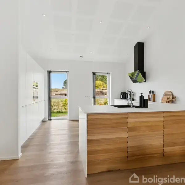 Et moderne køkken med en træbeklædt ø. Kaffemaskine og køkkenredskaber står på bordet. Væggen har to vinduer, der lukker lys ind. En vægur hænger over bordpladen.