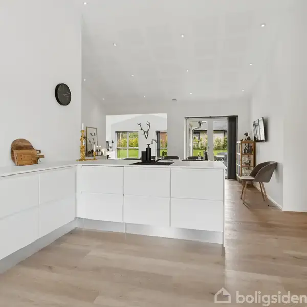 Lyst køkken med hvide skabe, sort emhætte og træbordplade. Rummet åbner op til en stue med store glasdøre, der fører til en terrasse. Væggene er hvide.