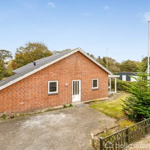 En rød murstensvilla står stille på en lille, grusbelagt gårdsplads omgivet af grønne buske og træer. Foran ses en dør med to vinduer, og himlen er klar.