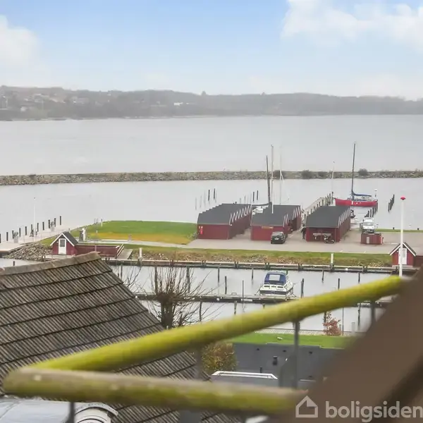 Set fra vindue mod havn med både i vandet, røde skure på kajen og roligt vand med bakker i horisonten. Hustage og tagrender ses i forgrunden.