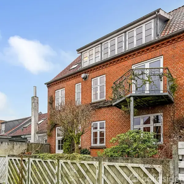 En rød murstensbygning med tre etager og en balkon. Bygningen er omgivet af et hegn og placeret i et kvarter med andre huse. Himmel med skyer ses i baggrunden.