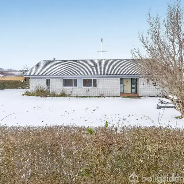 En enkeltstående træhus står i et snedækket landskab med en bar træ i forhaven. Huset er omkranset af en lav hæk, og der er blå himmel i baggrunden.
