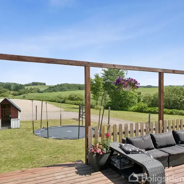 En udendørs terrasse med sort loungesofa set i træramme omgiver en gårdhave. En trampolin står på græsset, og en lille legehytte ses ved siden af i en åben, grøn landskab.