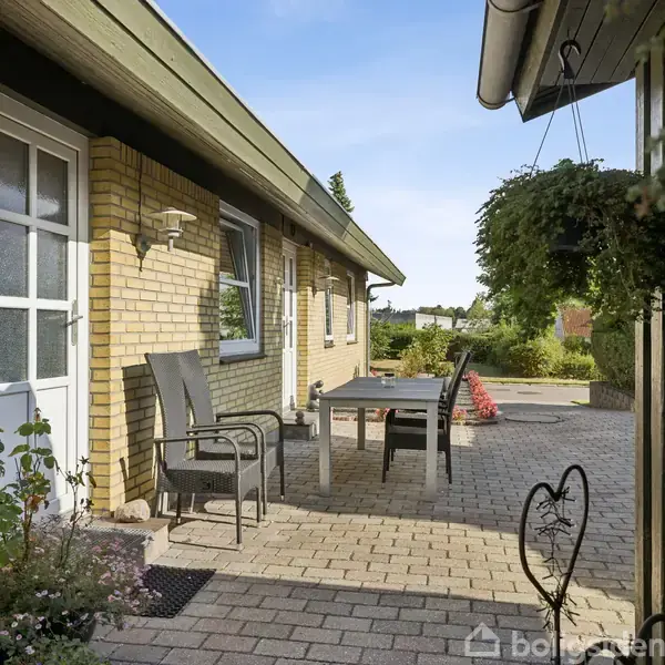Hus af gule mursten med udendørs terrasse, indeholdende bord og fire stole. Omkring er en flisebelagt gårdhave med planter og hængende potteplanter, omgivet af grønne områder og klart vejr.