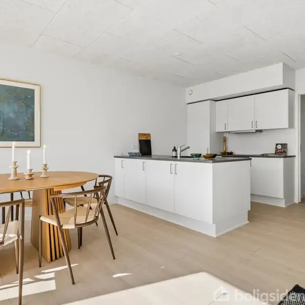 Spisebord med fire stole og to lysestager står nær en moderne, lys køkkenø i et åbent, minimalistisk rum. Et maleri hænger på den hvide væg i baggrunden.