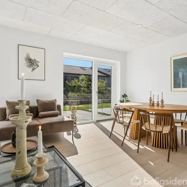 En moderne stue med to sofaer, hvorpå der ligger puder, står omkring et glasbord med tændte stearinlys. Rundt spisebord med stole placeret nær store vinduer med udsigt til haven.