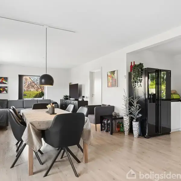 Et moderne køkken-alrum med spisebord og sorte stole i midten, to sofagrupper bagerst og et sort køleskab til højre. Lysindfald fra store vinduer skaber en luftig atmosfære.