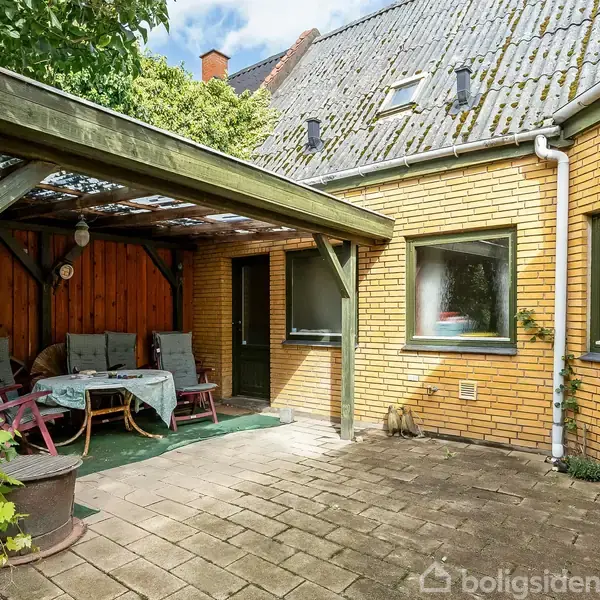 Terrasse med træoverdækning og havesæt i en baggård af gulstenshus. Omkringliggende vegetation inkluderer klatreplanter langs siderne. Vinduer på husets facade giver dagslys ind i rummet.