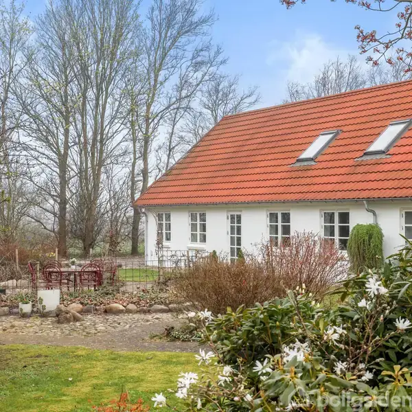 Hvidt hus med rødt tegltag og ovenlysvinduer står i en frodig have. Grussti og bede med blomster omgiver huset. Træer kan ses i baggrunden under en blå himmel.