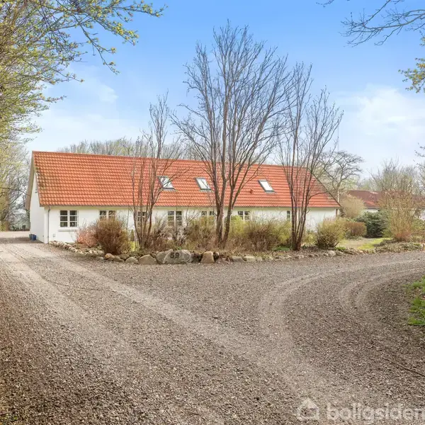 Hvidt hus med rødt tegltag står omgivet af træer langs en grusvej. Træerne har ingen blade, hvilket antyder forår eller efterår. Der er ingen synlig tekst i billedet.