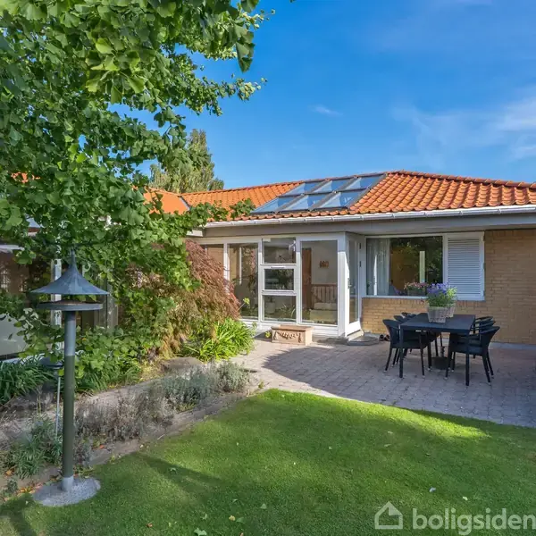 Et murstenshus med rødt tegltag omkranset af have med bord og stole på en flisebelagt terrasse. Grønne træer og buske omkranser området under en klar blå himmel.
