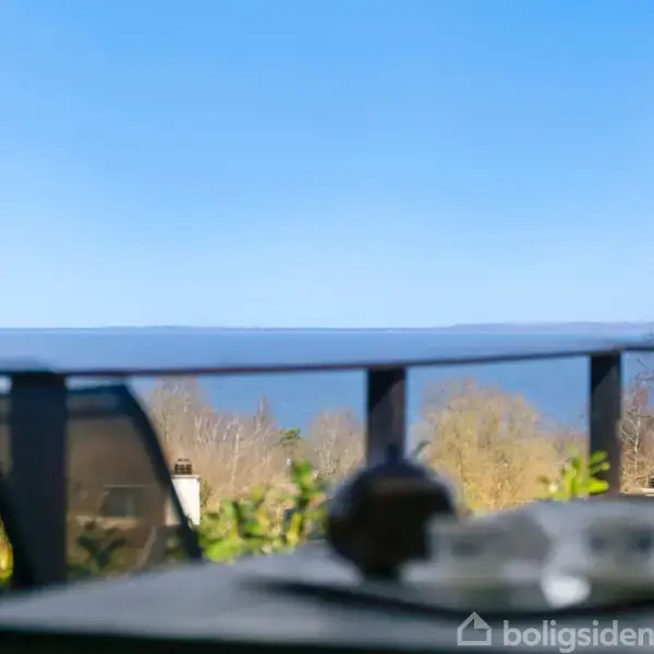 Terrassebord med tepotte og kopper står stille, omgivet af natur med træer og udsigt over en klar blå himmel og en rolig kystlinje i baggrunden.