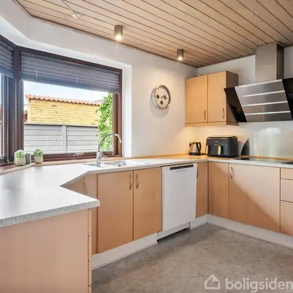 Et lyst køkken med træloft, beige skabe og moderne apparater, herunder en emhætte. Store vinduer giver udsigt til en terrasse med hegn. Vægur hænger over køkkenbordet.