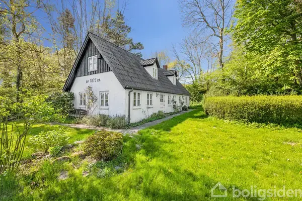 Et hvidt bindingsværkshus med sort tag står omgivet af frodig grøn vegetation og træer. Tekst på huset viser "Anno 1875". Huset ligger i en solrig have.