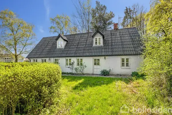 Hus med sort tegltag og hvide vinduer står i sollys. Det omkranses af velplejet græsplæne og grønne buske, omgivet af træer i en frodig have.