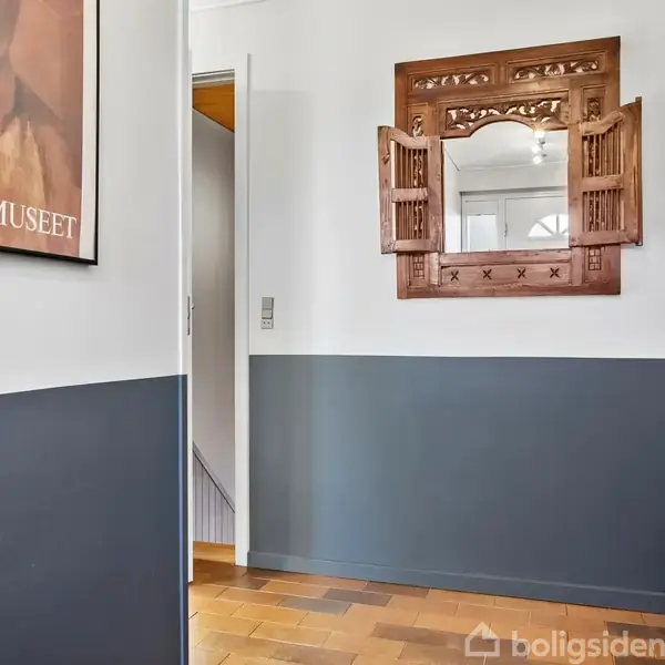 Et vægspejl med træramme hænger på en todelt væg; nedenfor står en brandslukker. En plakat med teksten "BLIXEN MUSEET" ses i venstre side.