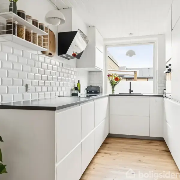 Et moderne køkken med hvide skabe og sorte bordplader. Der er en plante i nærheden og en opslagstavle på væggen. Et vindue giver lys ved vasken.