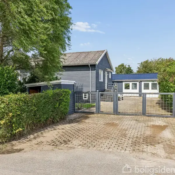 Gråt hus med træbeklædning står stille i en rolig villavej, omkranset af grønne buske og et hegn. En indkørsel med brosten fører til en metalport ved indgangen.