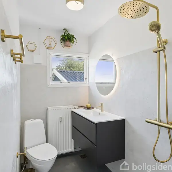 Et moderne badeværelse med en bruser i guld, en hvid toiletkumme, en sort håndvask med spejl og planter i gyldne vægdekorationer. Vindue og radiator til venstre.