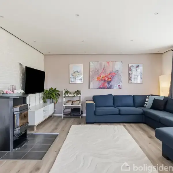 Stue med blå hjørnesofa står ved siden af store vinduer. Væggene har kunstbilleder. Der er et fladskærms-tv ved siden af brændeovn, og kurv står på gulvet.