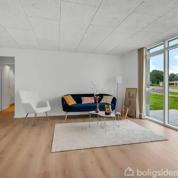 En stue med en blå sofa og hvid stol står på et lyst trægulv. To små runde borde er foran sofaen. Store vinduer giver udsigt til en have.