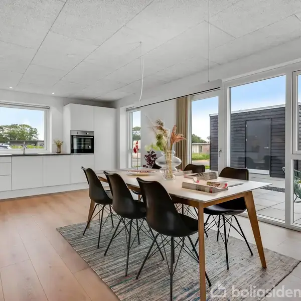 Spisebord med sorte stole står på et tæppe i et lyst, moderne køkken-alrum med store vinduer. Udsigt til en terrasse og en græsplæne udenfor.