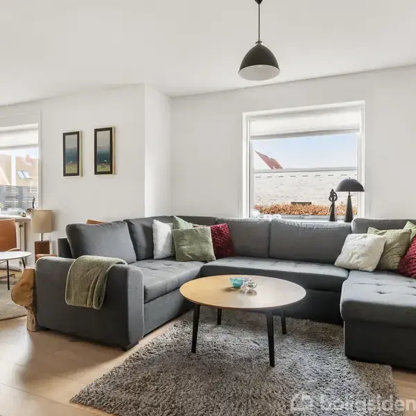 Stor, grå hjørnesofa med puder, placeret i veloplyst stue med trægulv, hvide vægge og to vinduer. Rundt sofabord med dekorative genstande foran sofaen. Spiseplads i baggrunden.