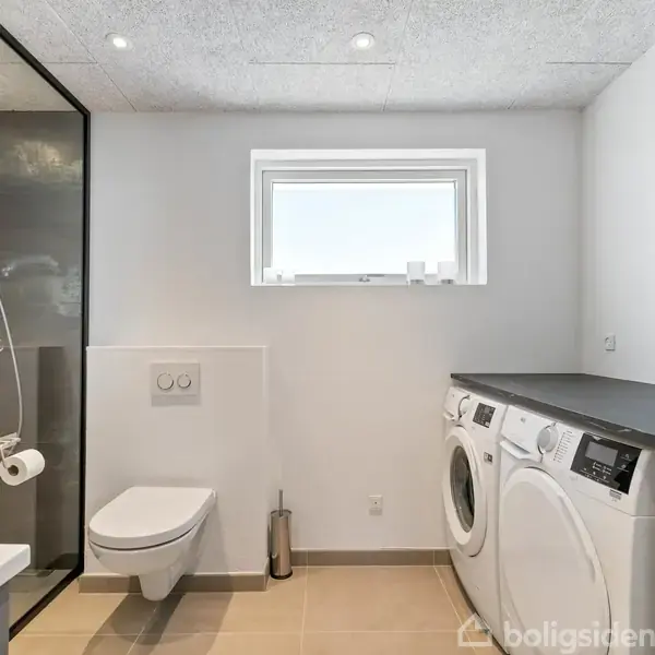 Elegant badeværelse med væghængt toilet, glasbrusekabine og vaskesøjle med vaskemaskine og tørretumbler. Rummet er lyst og moderne med et lille vindue og gulve af beige fliser.