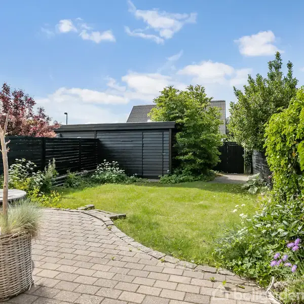 En have med græsplæne og planter omkranset af et sort hegn. Til venstre står en kurv med planter og en træbænk, mens himlen er blå med skyer.