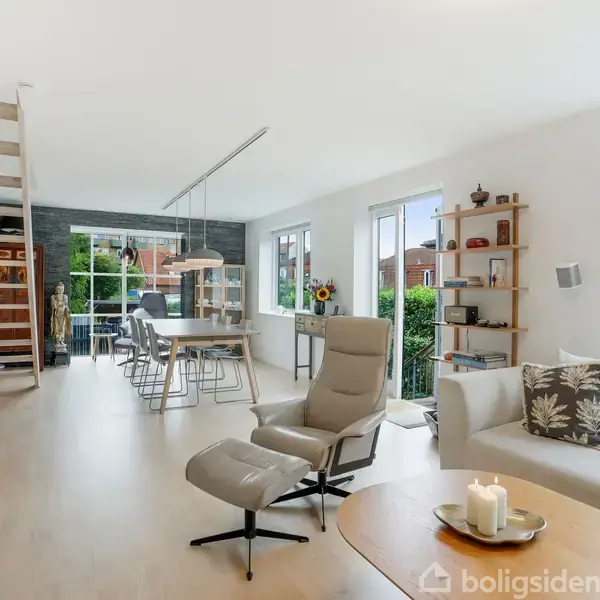 Stue med moderne indretning. Sofa og stol står ved et sofabord med stearinlys. Trappe fører til en overetage. Store vinduer giver udsigt til haven, spisestue i baggrunden.