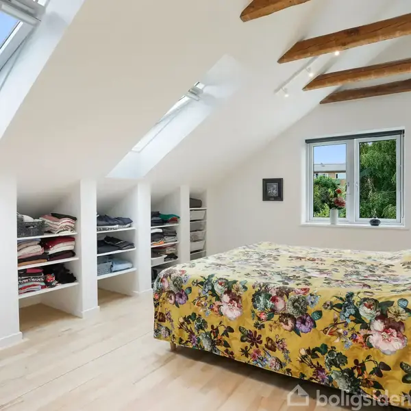 Soveværelset har en seng med et blomstret tæppe, placeret under skrå lofter med ovenlysvinduer. Der er åbne hylder til tøj langs væggen, og en enkelt væghængt lampe.