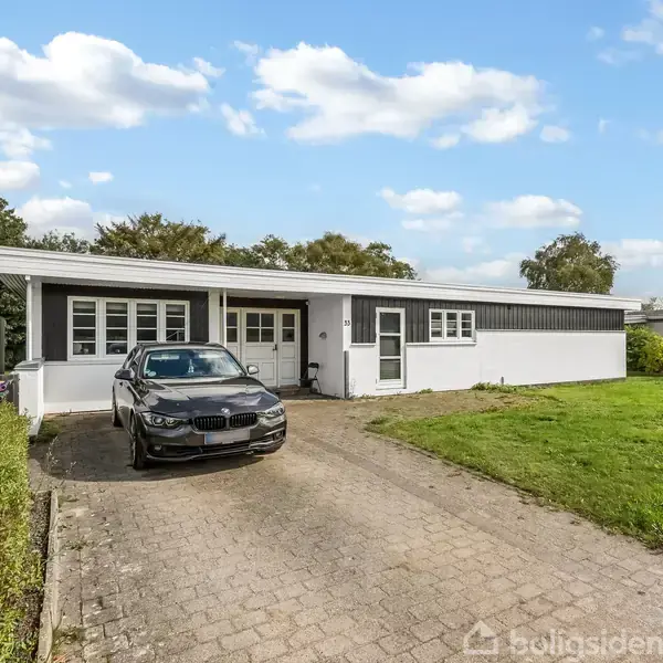 En sort bil holder parkeret i indkørslen foran en hvid bungalow med fladt tag, omgivet af en velplejet have og grønne træer i baggrunden.