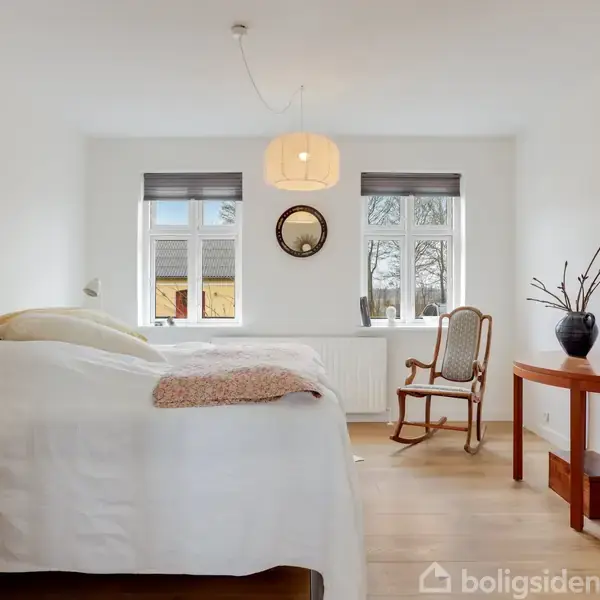 Soveværelse med stor seng til venstre, en gyngestol for enden, og et lille bord med vase til højre. Værelset har to vinduer og en loftslampe.