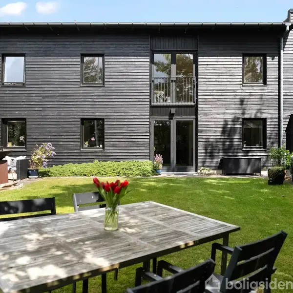 En moderne sorte træfacadebygning står stille omgivet af en grøn have. Foran huset er et udendørs bord med seks stole, og en vase med røde tulipaner står på bordet.