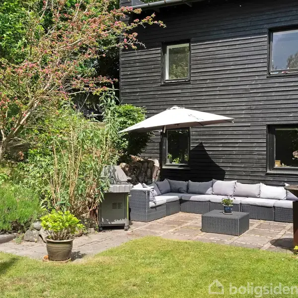En sortmalet træhusvæg danner baggrund for en udendørs terrasse med en grå hjørnesofa under en parasol. Planter og en grill pryder den solrige have.