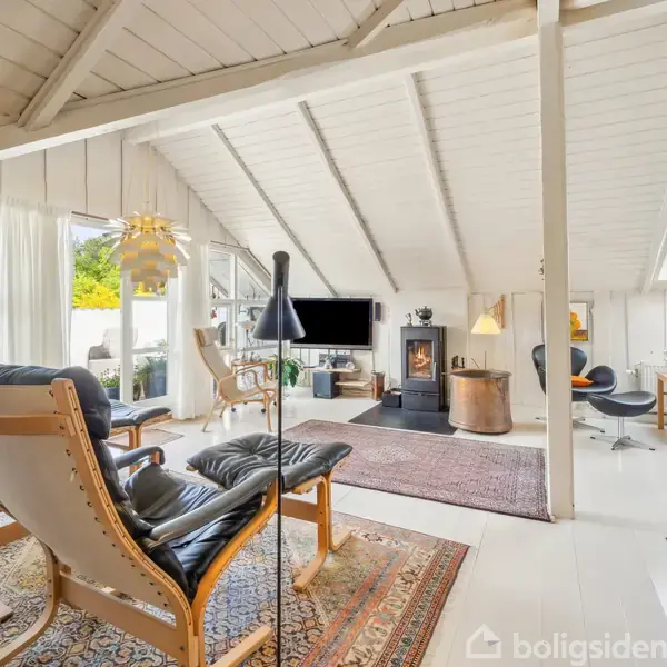 Stue med moderne lænestole, pejs og fladskærms-TV under et skråt loft. Rummets store vinduer giver udsigt til en grøn have. Indretningen omfatter tæpper og belysning.