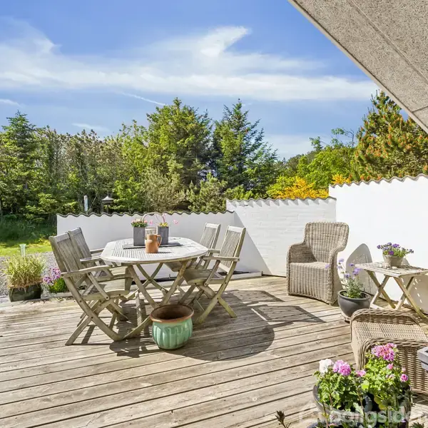 Terrasse med træmøbler inkluderer et rundt bord og stole, omgivet af planter i krukker. Hvid mur til højre, og grøn have med buske og træer i baggrunden under en klar blå himmel.