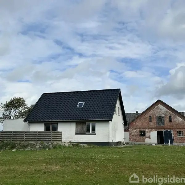 Et hvidt hus med sort tag står foran en stor græsplæne. Ved siden af er en ældre murstensbygning. Skyer fylder himlen i baggrunden.