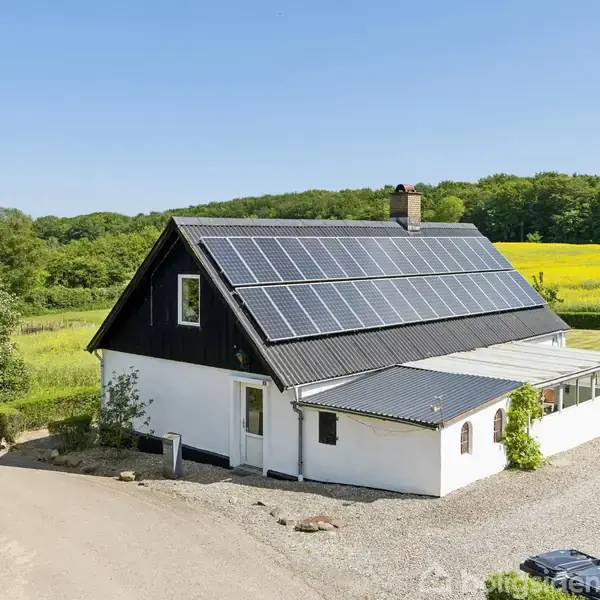 En hvidmalet landejendom med solpaneler på taget står ved en grusvej. Omgivet af en grøn eng og en skov i baggrunden, under en klar blå himmel.