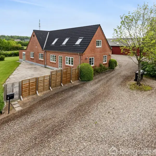 Et rødt murstenshus med sort tag har flere vinduer og en grusvej foran. Omkring huset er en frodig have med træer og buske.