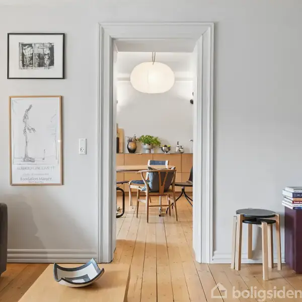 Stue med grå sofa, lampeskærme og plakat på væggen. Åben dør fører til spisestue med rund lampe, bord og stole, omgivet af lys træindretning. bøger og skammel til højre.