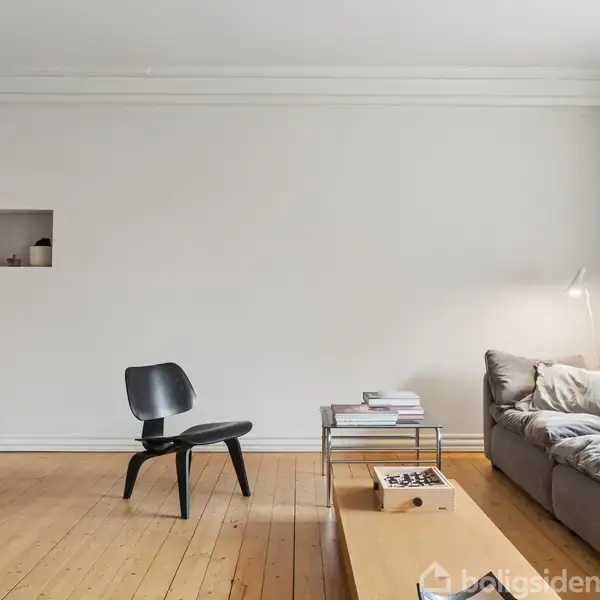 Et moderne, minimalistisk stueinteriør med en grå sofa og en sort stol på et lyst trægulv. En væglampe lyser på sofaen, og der er bøger på et lavt bord.