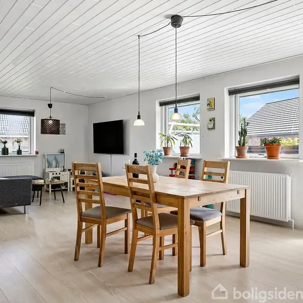 Spisebord med fire træstole står i en lys spisestue med træloft. Væggen har tre vinduer med planter. Sofa og tv ses i baggrunden. Ingen tekst er til stede.