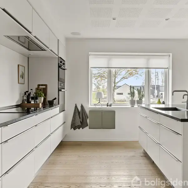 Moderne køkken med hvide skabe og sorte bordplader, arrangeret omkring en stor central ø. Et vindue bringer naturligt lys ind. Gulvet er af lyst træ. Ingen tekst.