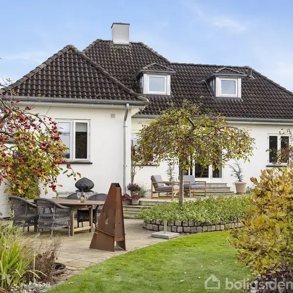 Hus med hvid facade og mørkt tag står i en have med stenbelagt terrasse, træer og grønne buske; havemøbler og pejs på terrassen. Skyfri himmel i baggrunden.
