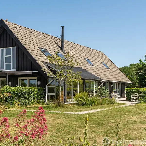 Et sortmalet hus med skråt tag står roligt i en frodig have med græs, blomster og træer. En sti fører til en udendørs terrasse med havemøbler.