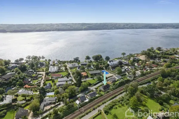 Hus markeret med blå pil tæt på jernbanespor, omgivet af villakvarter med træer og haver, beliggende ved en bred sø.