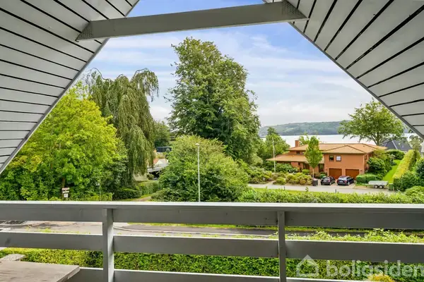Balkon med hvidt rækværk og skråt tag giver udsigt over en frodig have og et boligområde. I baggrunden ses et stort træ samt et rødt hus med parkerede biler.