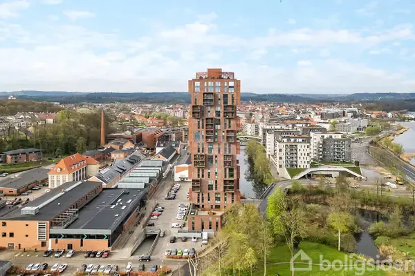 Høj bygning står stille i byområde omgivet af mindre bygninger, en flod og grønne træer. Skyskraberens moderne design skiller sig ud mod den bymæssige baggrund.