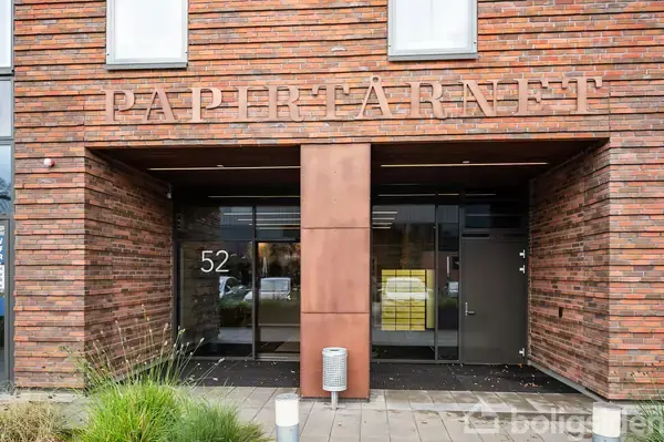 Bygningen har en glasdør indgang med teksten "PAPIRTÅRNET" ovenfor. Husnummer 52 er ved siden af døren i murstensbygningen med græs og planter foran.